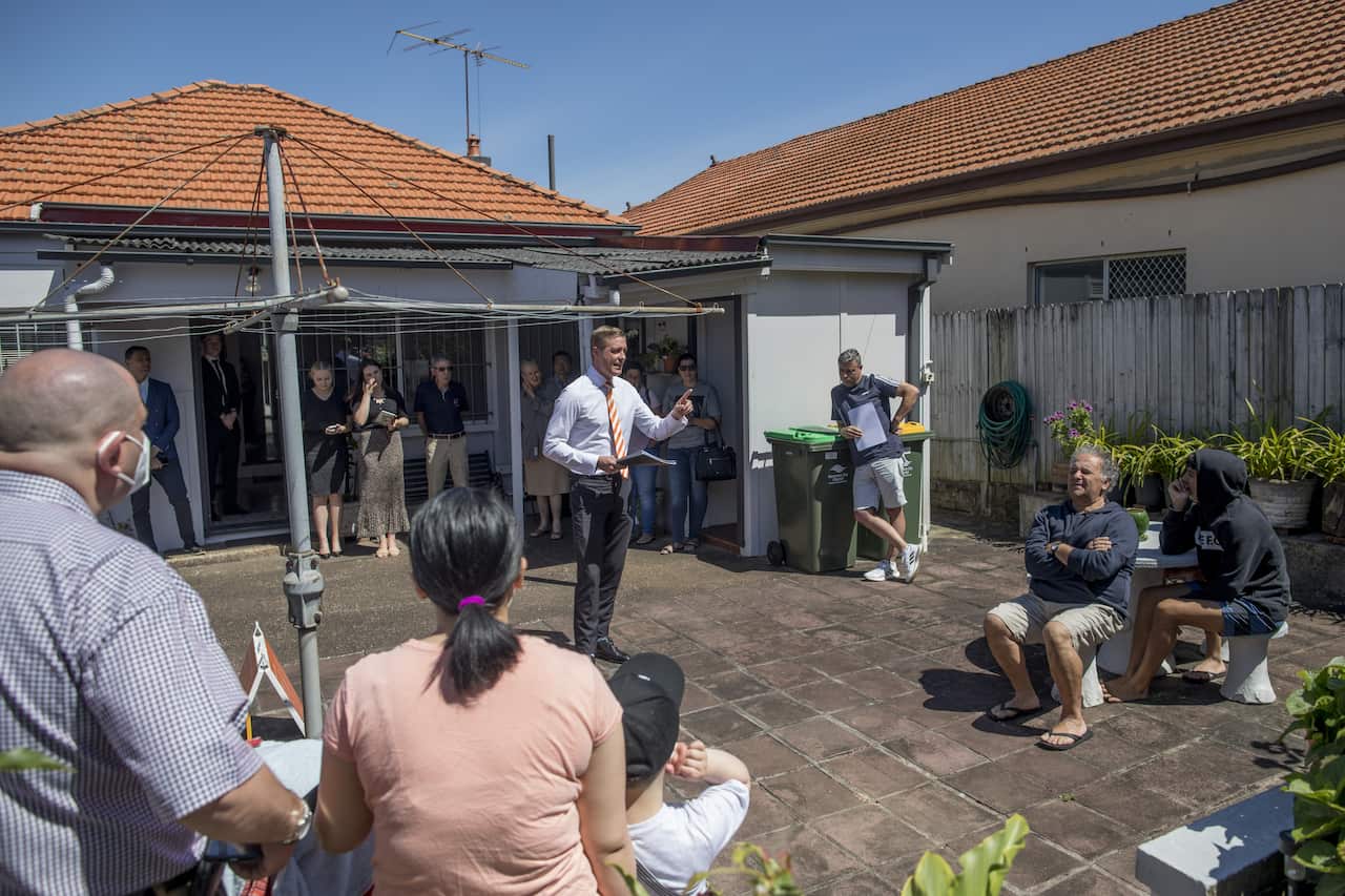 An auctioneer holding paperwork stands in the paved backyard of a brick home with a rotary clothesline, calling bids to a group of prospective buyers seated and standing around the patio.
