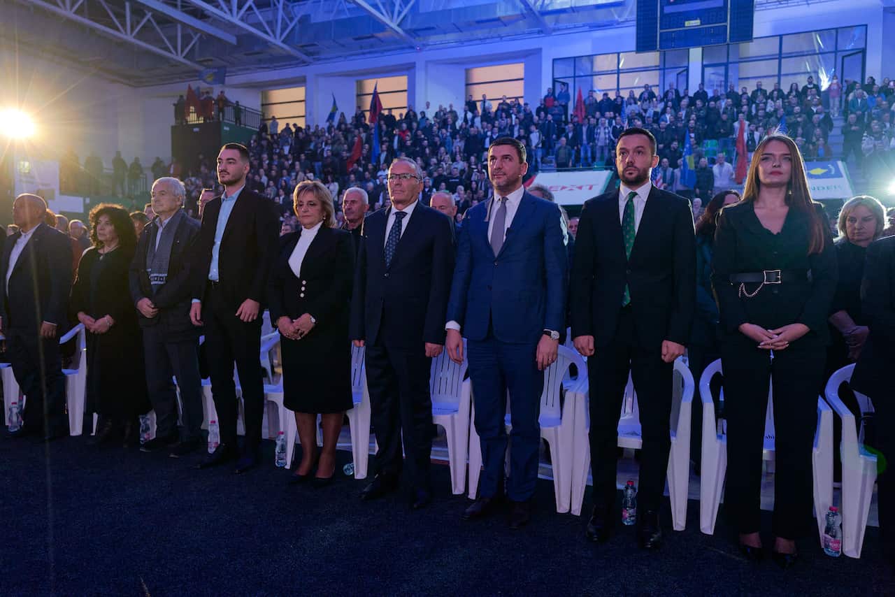 A line of officials and political figures stand at attention during a ceremony or event in a crowded indoor arena, with spectators and flags visible in the background.