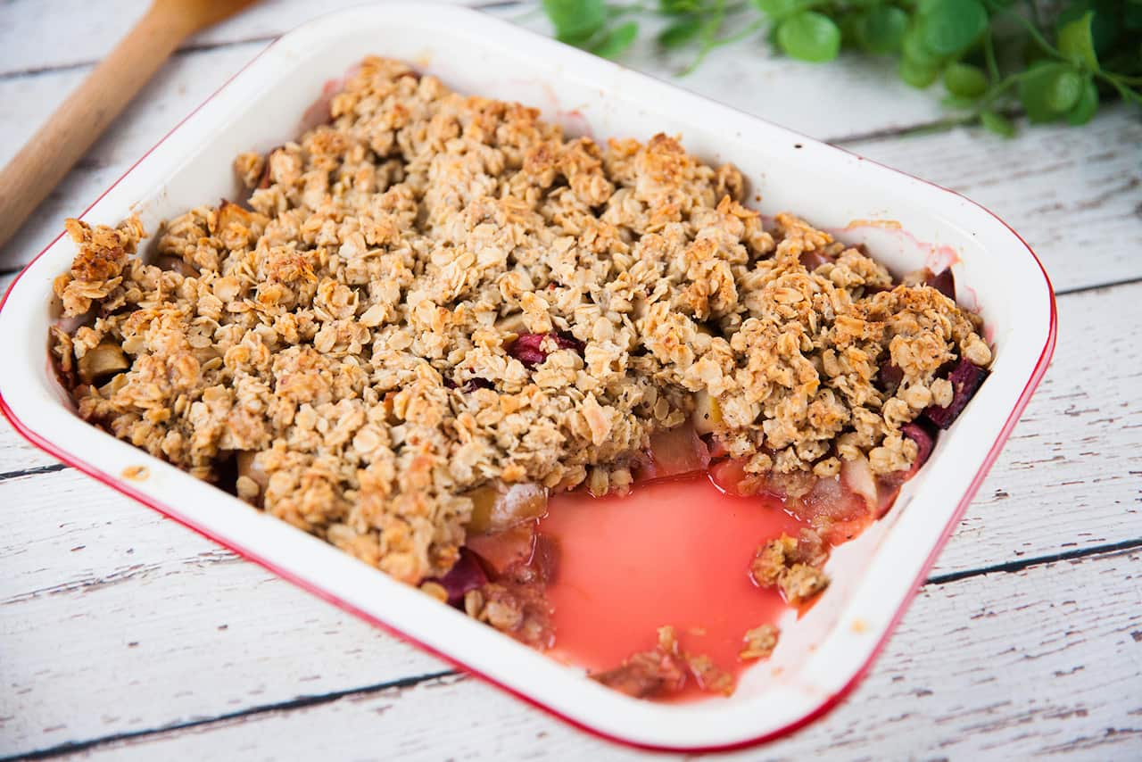 An old-style baking tray sits on a wooden surface. A portion of the crumble has been removed, showing the pink-tinged fruit under the chunky crumble topping.