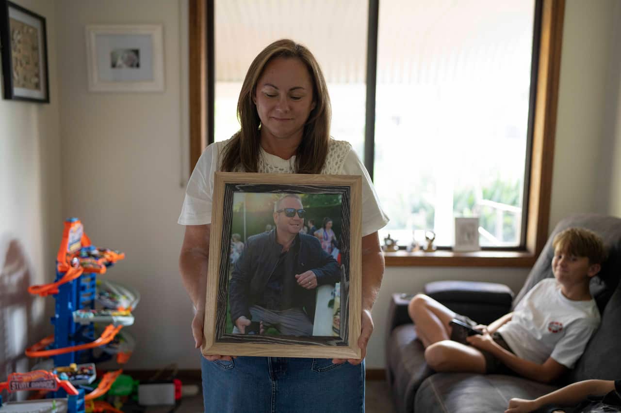 A middle-aged woman is standing in a small living room holding a framed photo of a man. A young boy is sitting on a couch behind her.