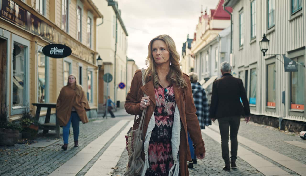 A woman with long hair, in a patterned dress, long coat and scarf, walks down a quiet street lined with shops.