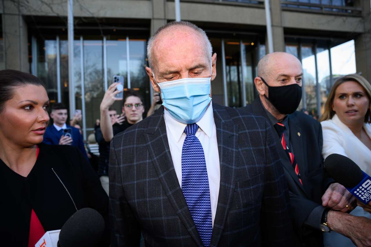 A man wearing a suit and a face masks leaves a court room flanked by media. 