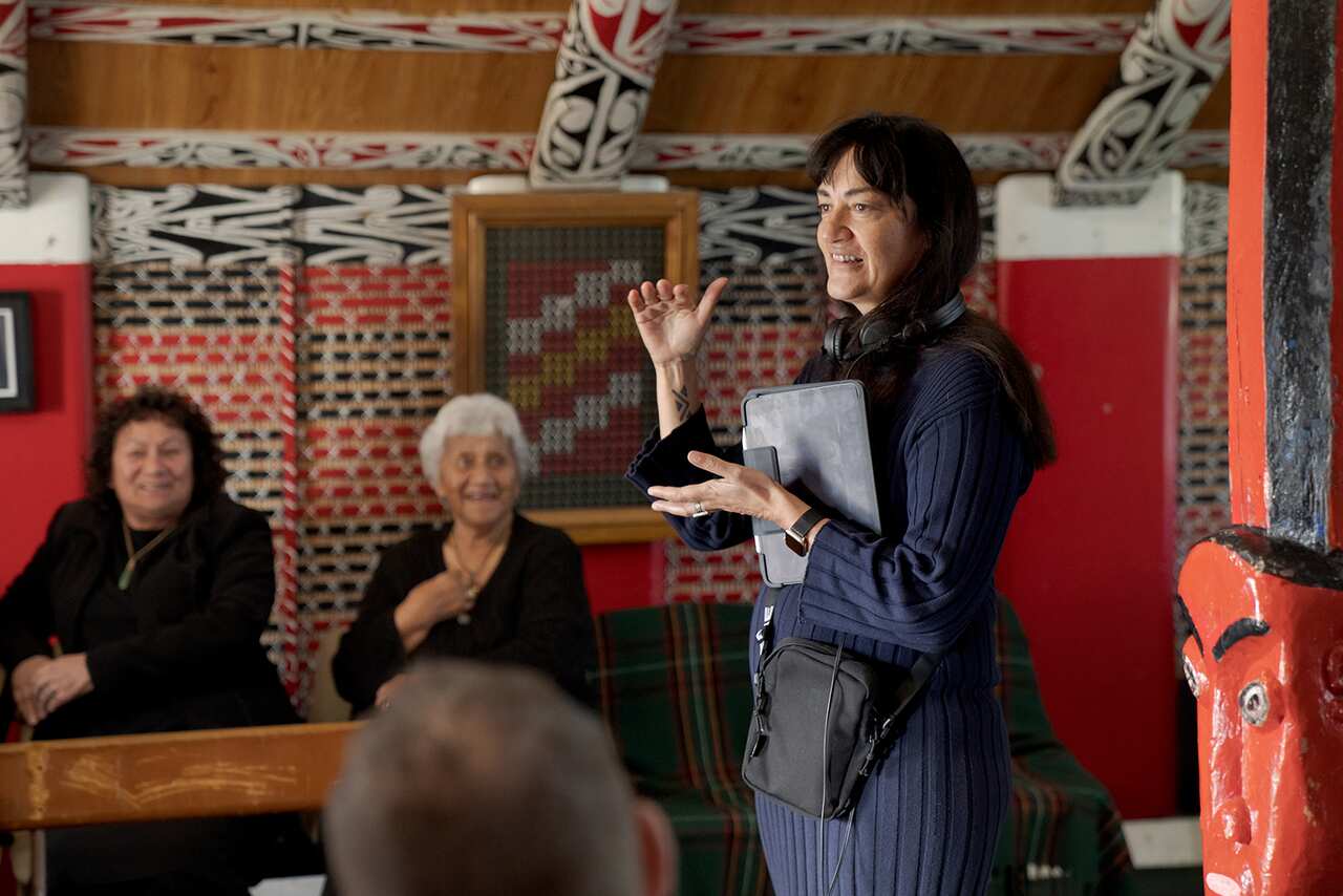A woman, gesturing with one hand and holding an iPad with the other, stands inside a room that appears to be decorated with Maori motifs. Two women, seated, watch her. 
