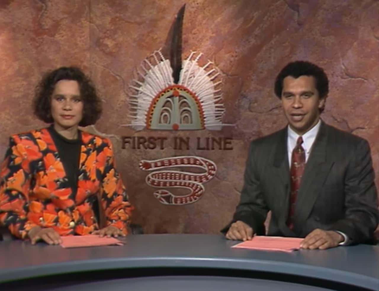 A female news anchor in an orange-and-black blazer sits next to a male anchor in a suit. There's an Indigenous logo with "First in Line" written at the bottom, behind them.