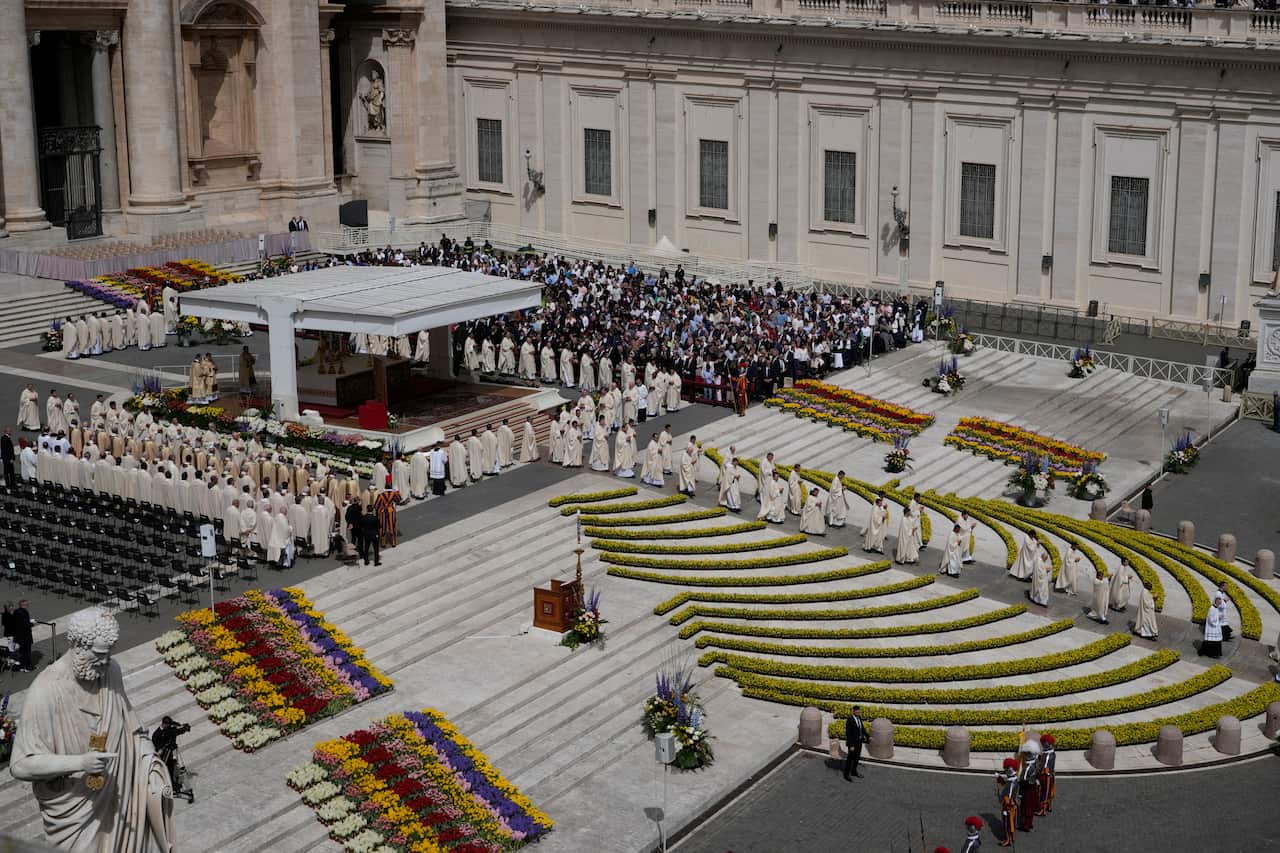 A photo from above shows a procession of white-robed clergymen and a small group of visitors in a historical European square. 