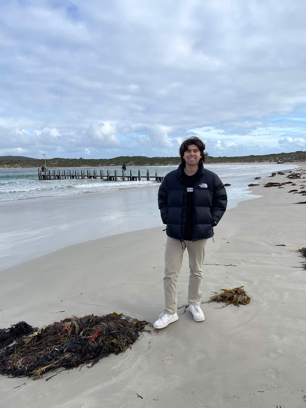 A man wearing a dark puffy parka, cream pants and white sandshoes stands on a beach.