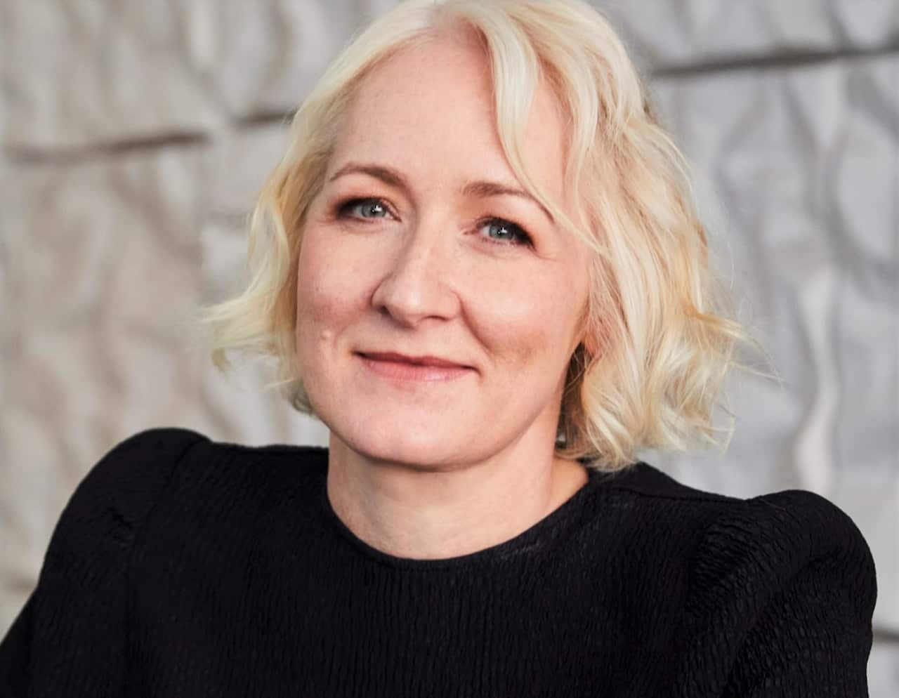 A blone haired woman in a black top smiles at camera.