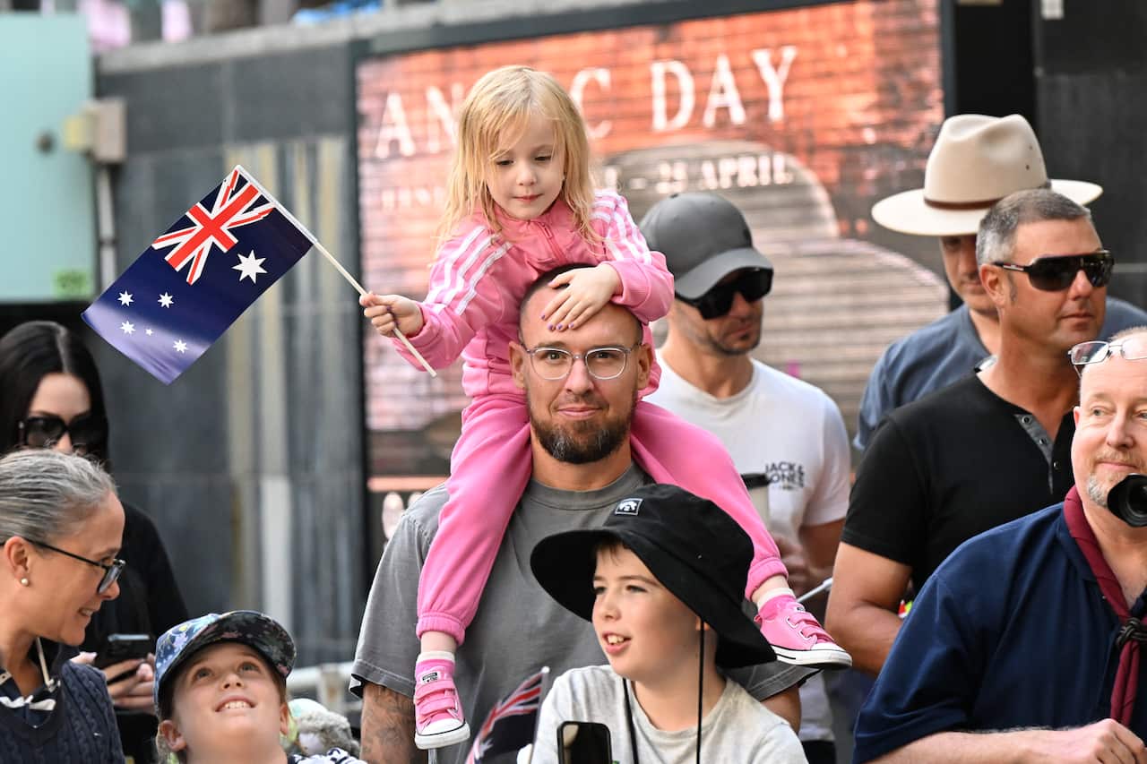 A group of people. A girl wearing a pink tracksuit and holding an Australian flag on a stick is sitting on a man's shoulders.