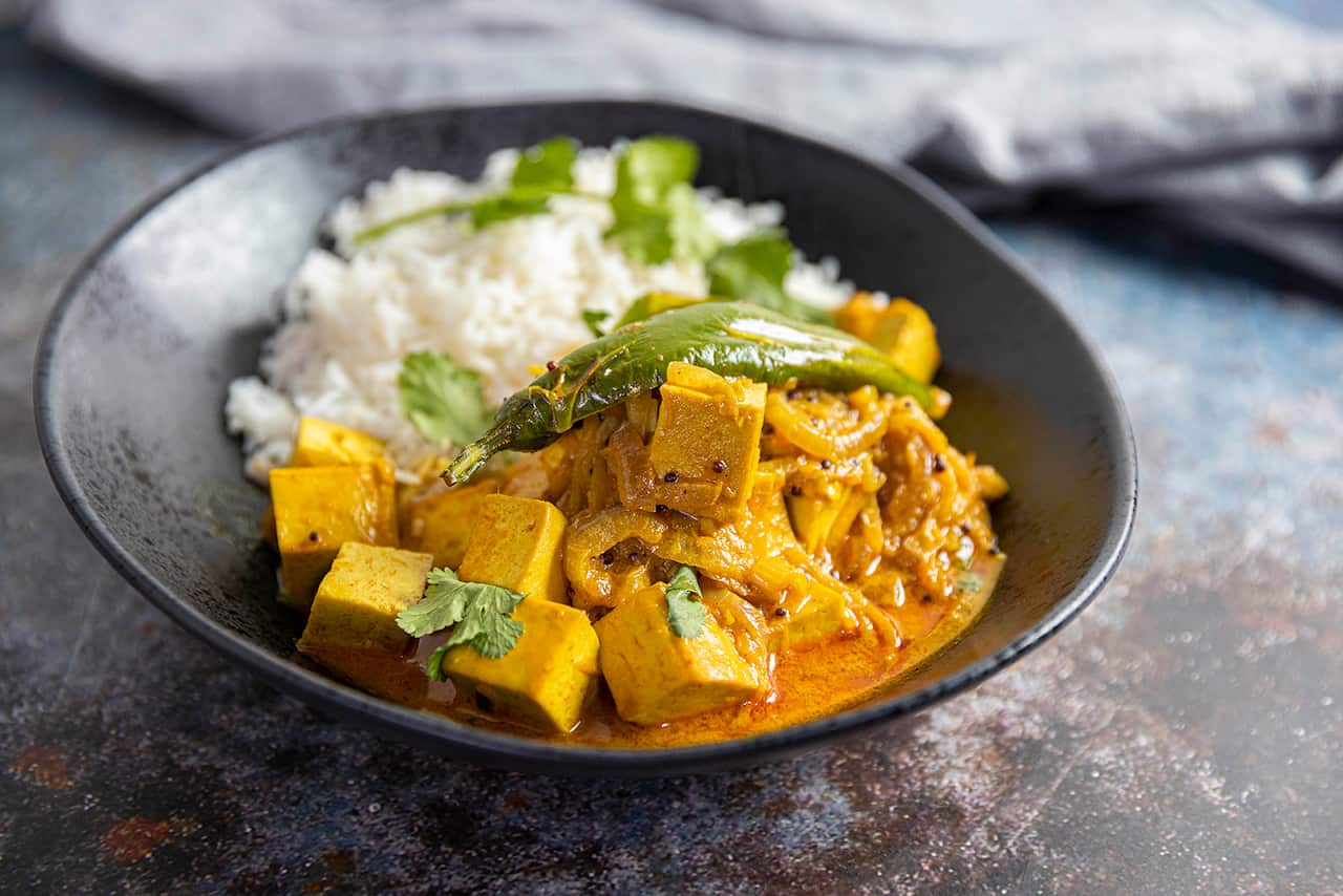 A vibrant yellow tofu curry and white rice sit in a dark, modern ceramic bowl. 