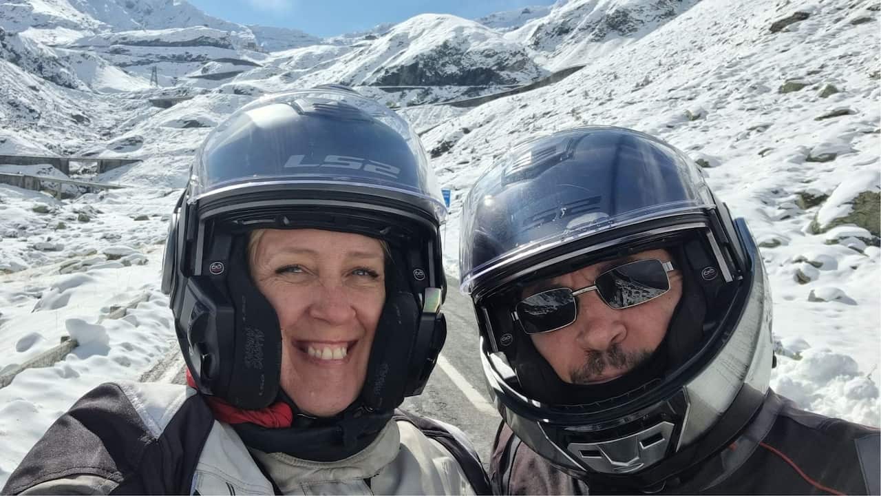 A middle-aged man and woman in motorbike gear and helmets take a selfie on a road in snowy mountains.