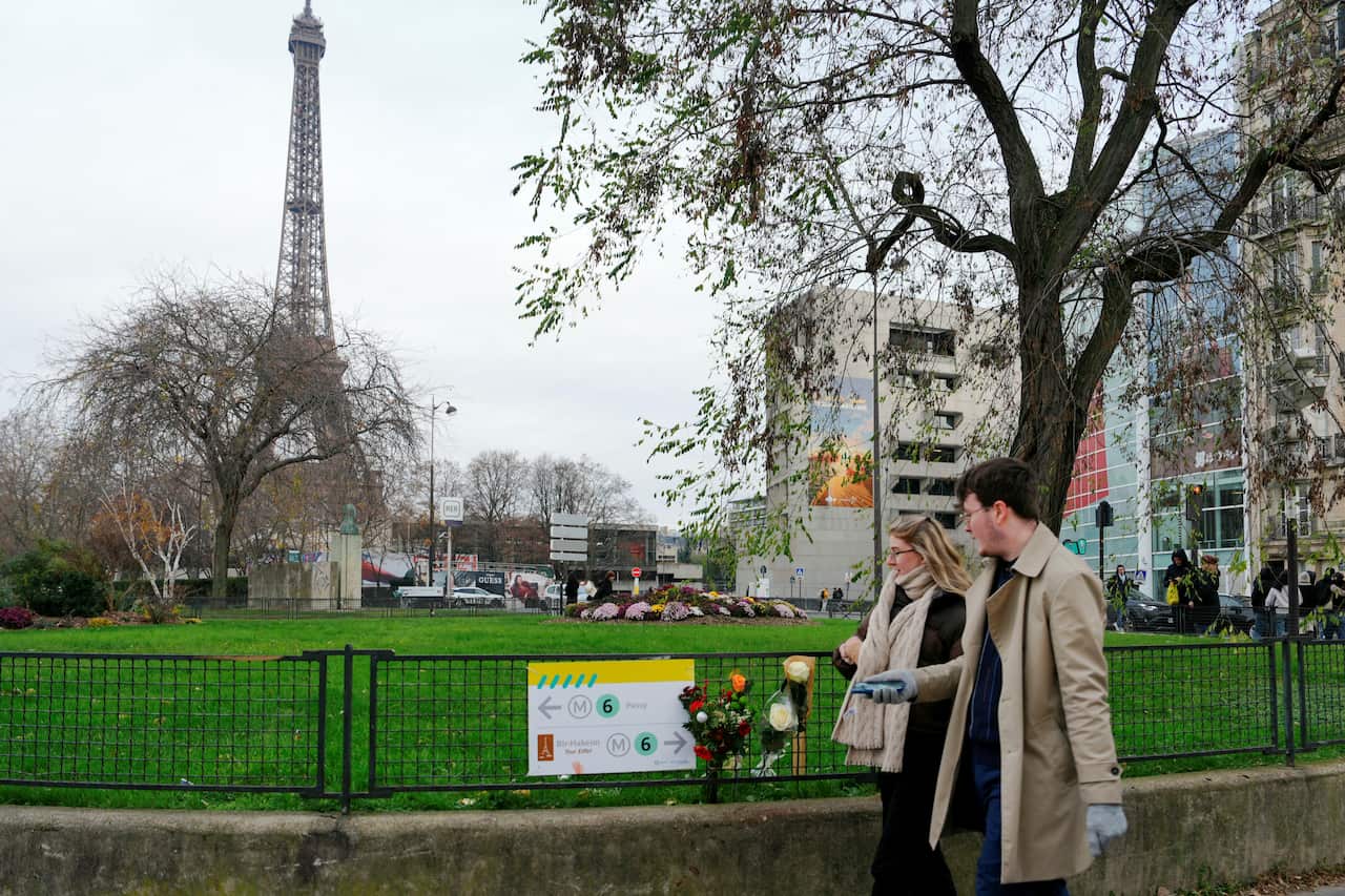 A man and a woman walk past flowers placed in a fence at the perimeter of a small park. The Eiffel Tower is in the background.