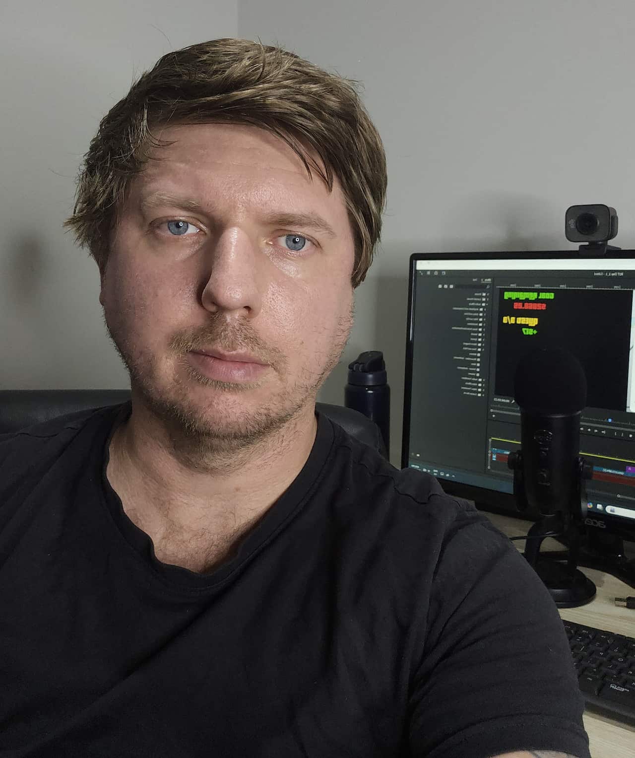 A man in a black T shirt looks at the camera with a neutral expression on his face.