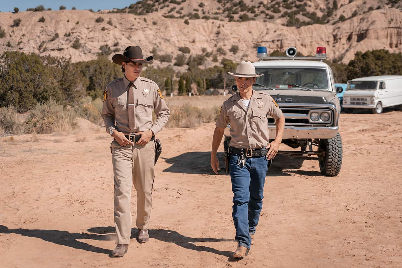 Two men in brown police uniforms and wide hats walk away from a four-wheel drive vehicle, across an expanse of red dirt.