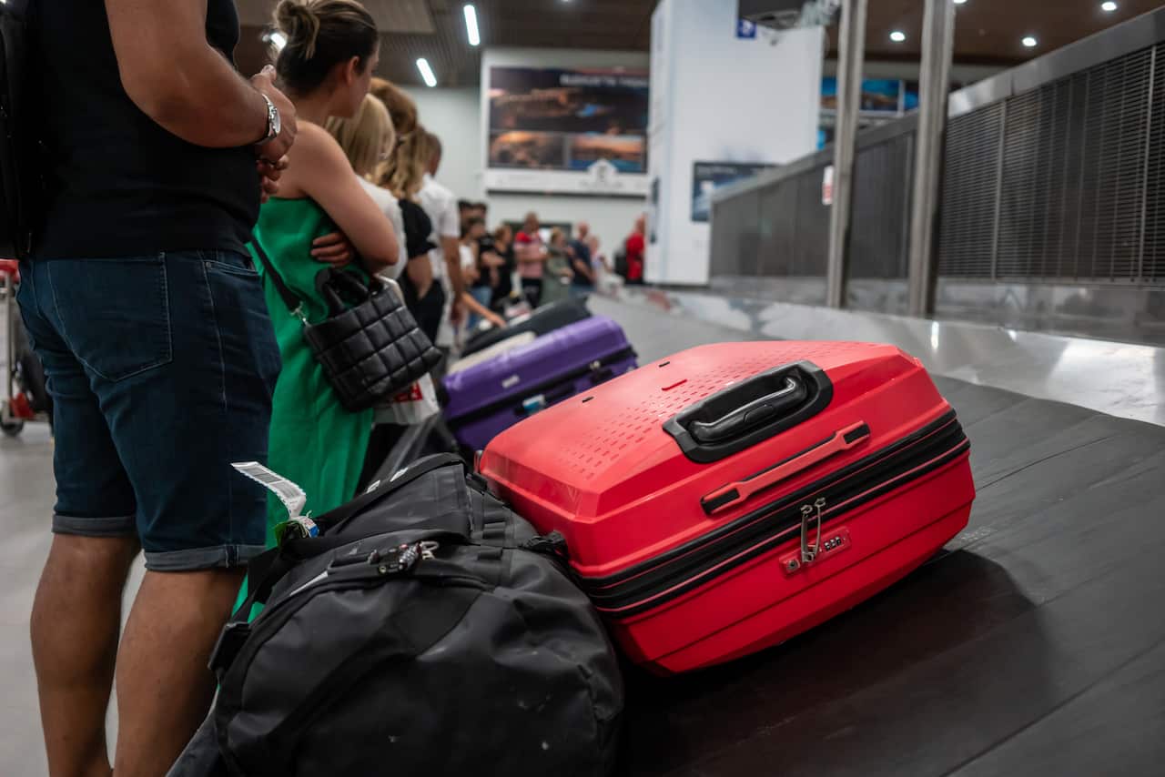 Passengers waiting for luggage on a carousel at an airport. There is a black bag, a red suitcase and a purple suitcase on it.