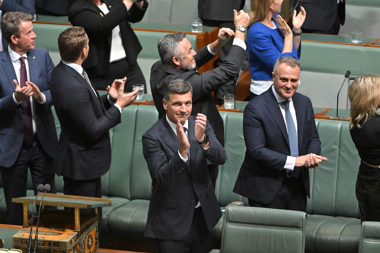 A man in a suit claps his hands and smiles while standing with other in the House of Represenatives.