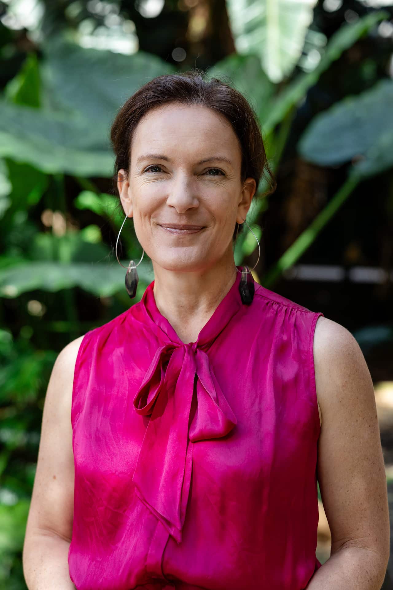 A woman with short, brown hair in a pink sleeveless top with a bow collar stands smiling in front of plants in a garden