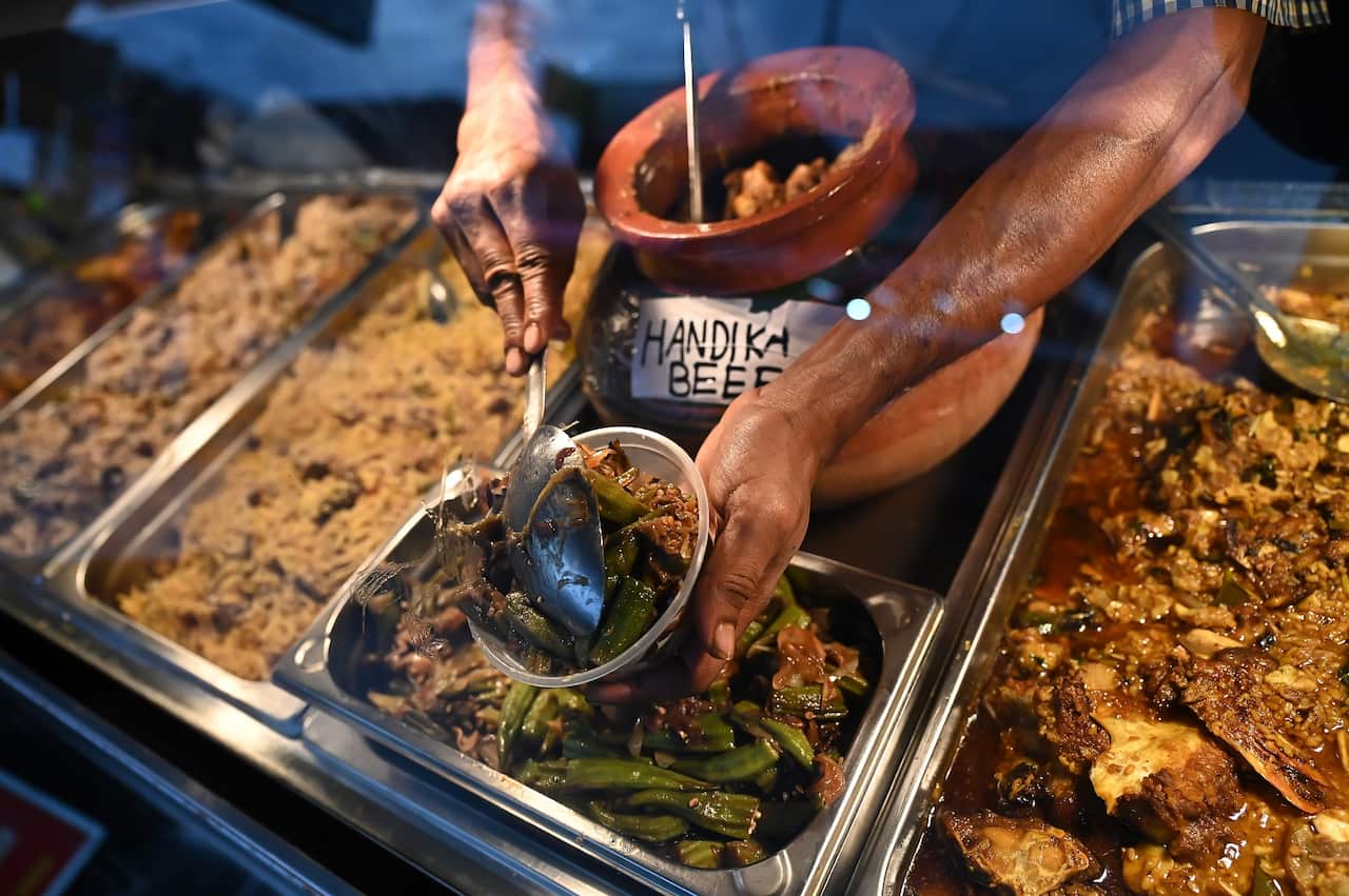 A food vendor uses a ladle to place food into a container from a counter filled with other dishes.