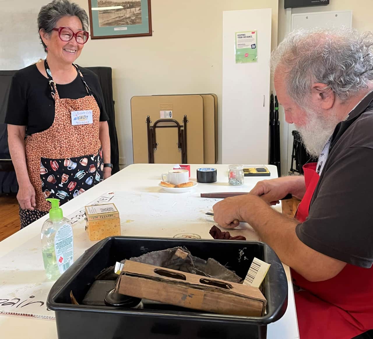 A woman in an apron stands next to table where a man sharpens a knife.