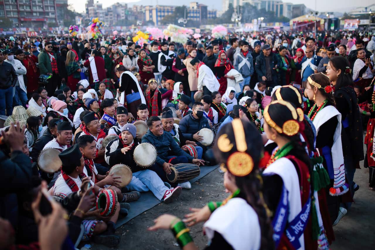 A group of people in traditional attires are gathered, some sitting and playing drums while others are standing and dancing as people watch.
