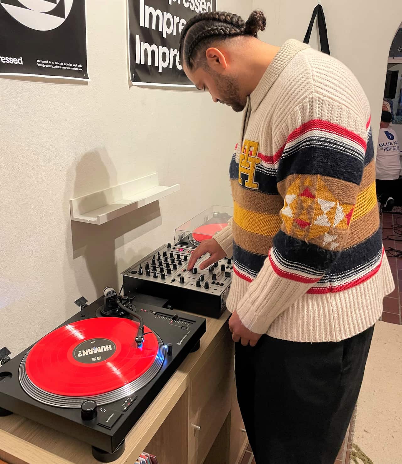 A man in a white jumper stands at a record player with a red record on the turn table.