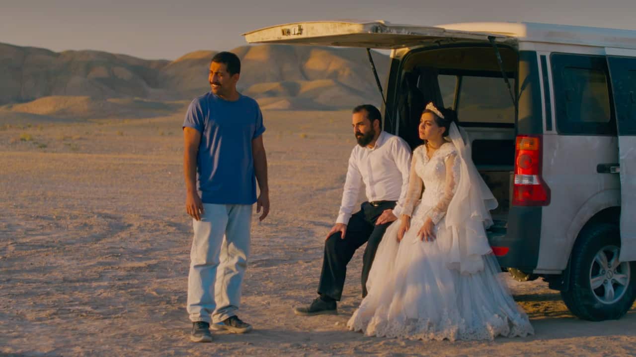 A bride and groom sit at the back of a van in a desert, as a man stands nearby under the setting sun.