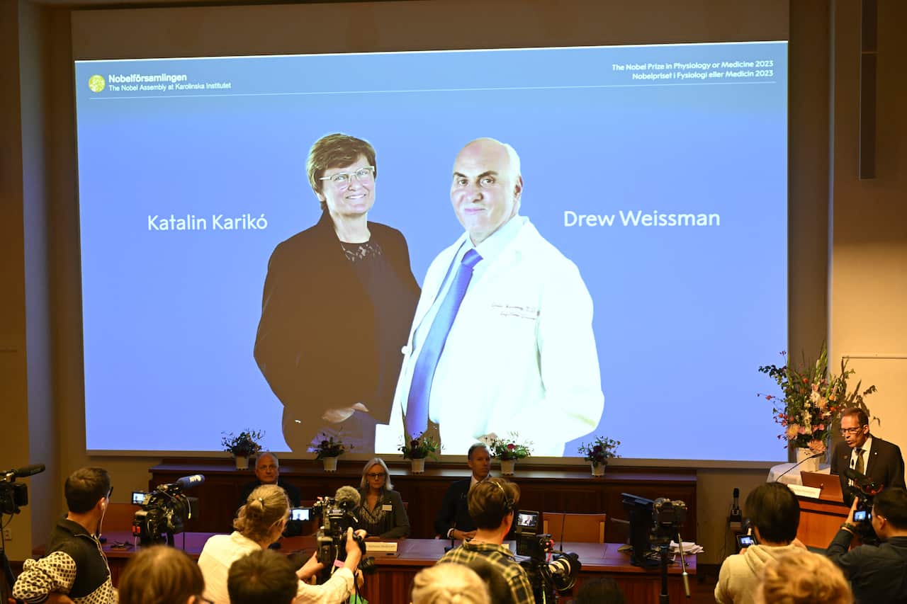 A man talks at a podium next to a table of people, with a blue screen showing Katalin Kariko and Drew Weissman behind them.