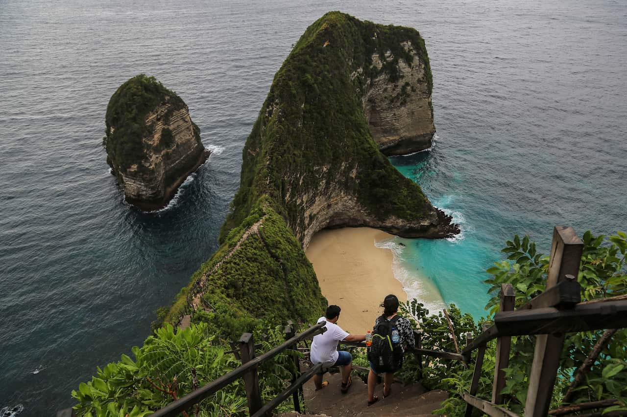 Tourists walk down a steep set of stairs to on a cliff down to a picturesque beach.