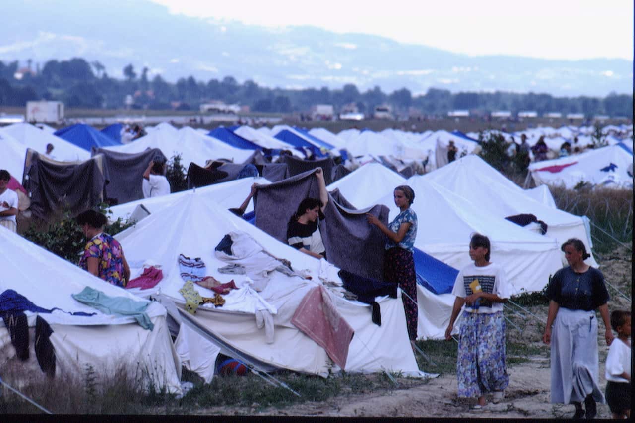 Tents as far as the eye can see, most white, some blue. Women are hanging sheets and walking between the tents. 
