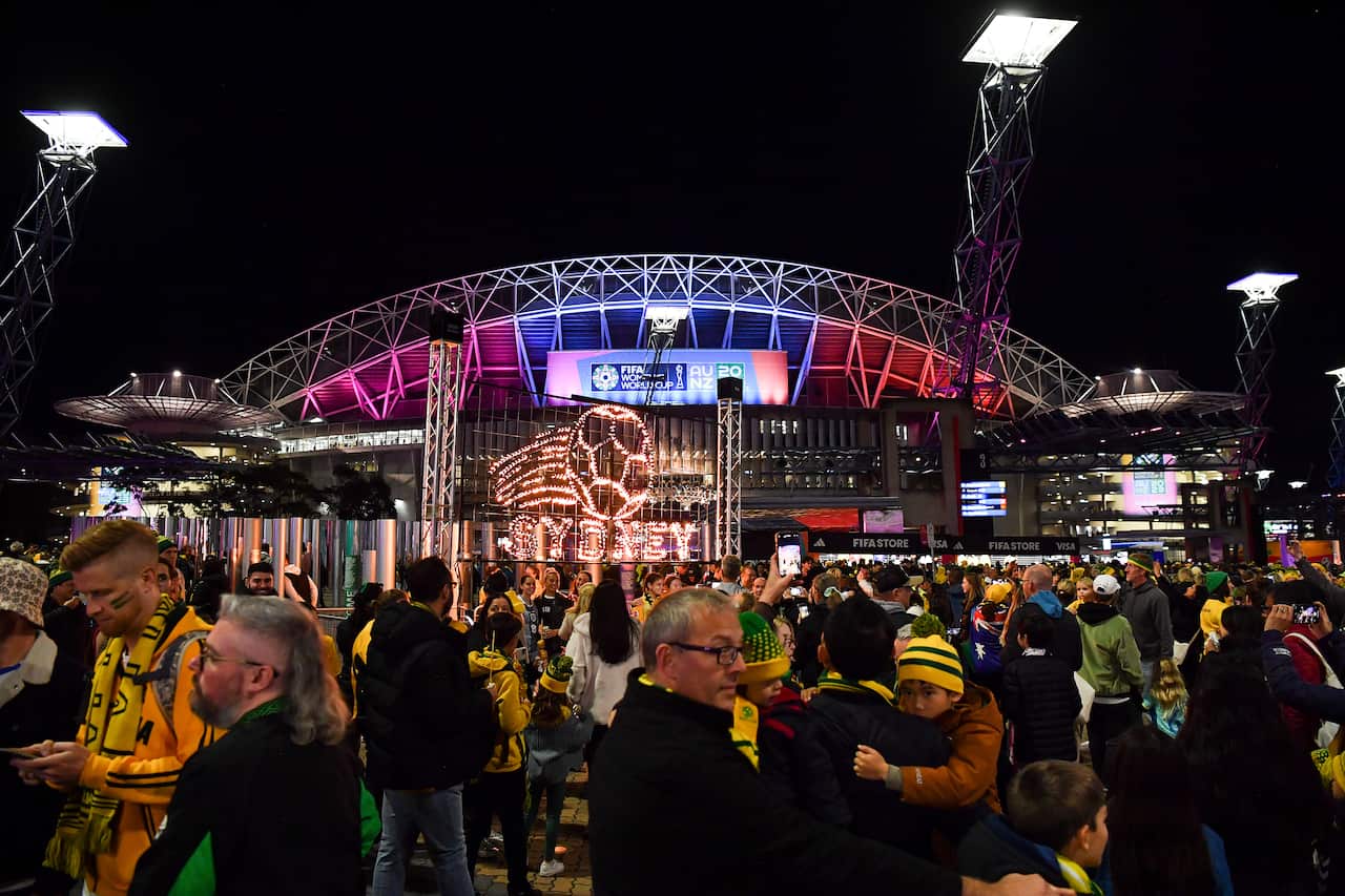 A crowd of people wearing yellow and green clothing accessories, standing outside of a stadium. 