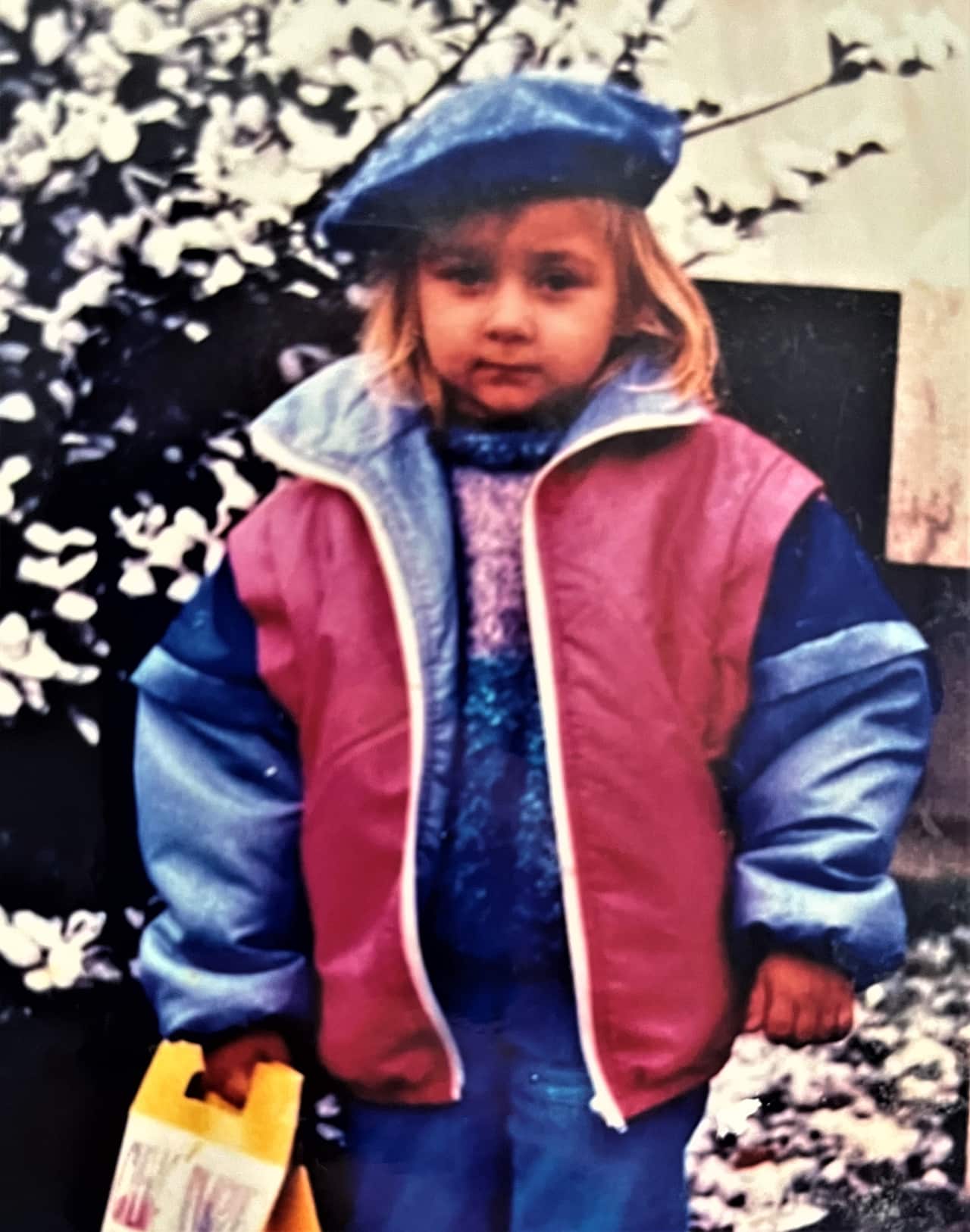 A child in a red and blue parka and a blue cap stands in a garden. 