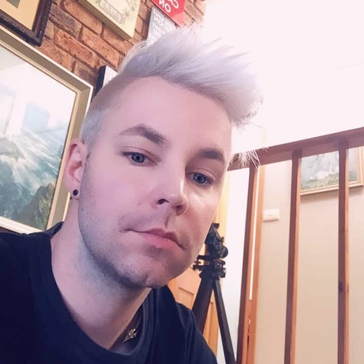 A man with bleached white hair stares down into the camera, a painting behind him on the wall