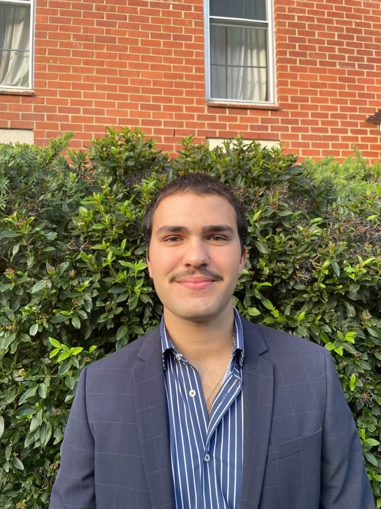 A man wearing a dark blue suit jacket and blue striped shirt poses in front of  a hedge. There is a brick building behind the hedge.