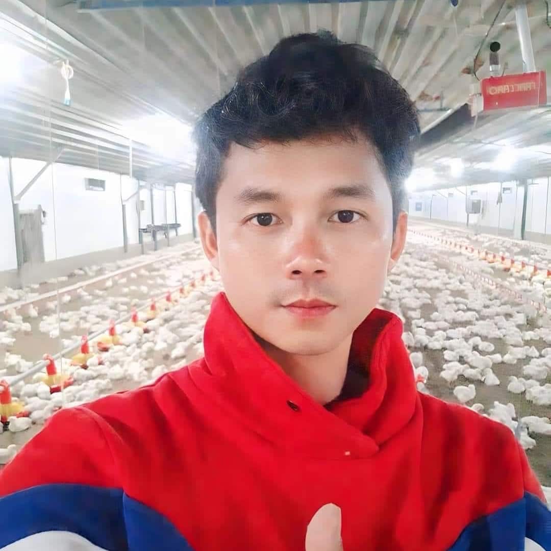 A young Thai man wearing a red pullover fleece in a shed with hundreds of chickens on the ground behind him.