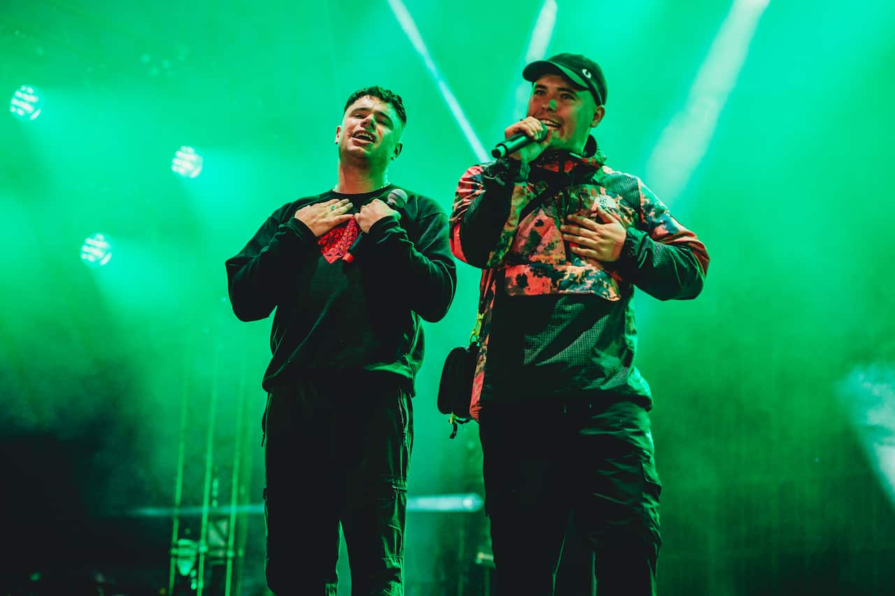 Two men singing on a stage. One is holding a microphone. There is green smoke in the background