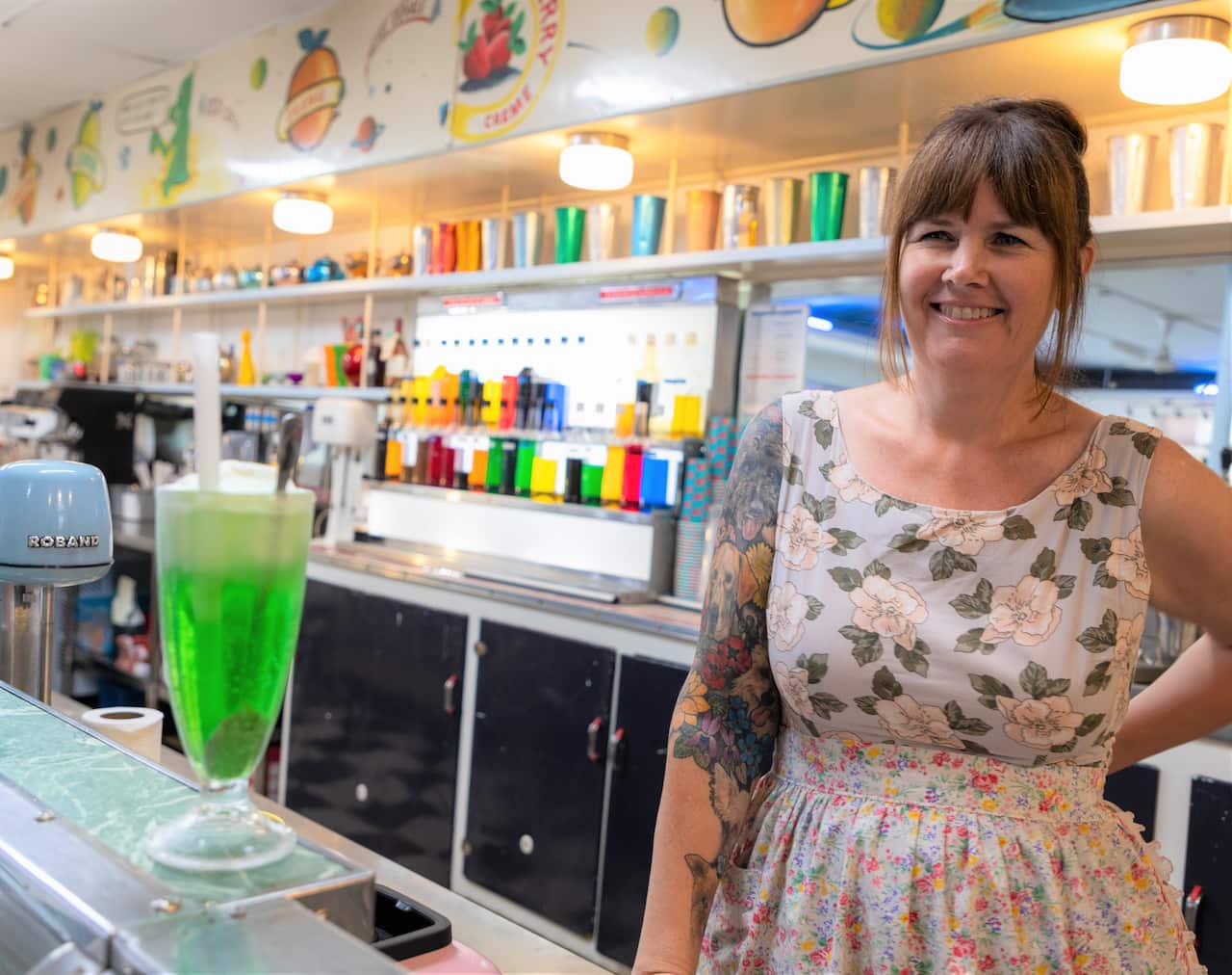 A woman in a floral top stands next to a green drink resting on a shop counter. 