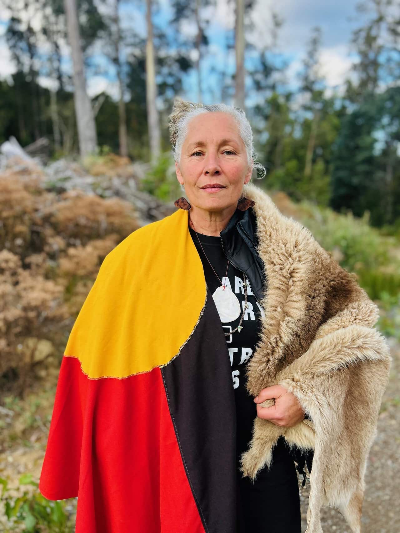 A woman standing outside posing for a photo. She has an Aboriginal flag draped over her right shoulder and an animal skin over her left shoulder.