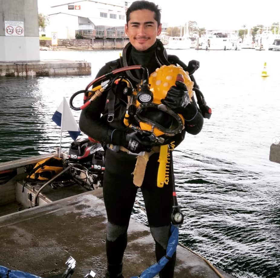 A commercial diver stands in his gear above the water and smiles.