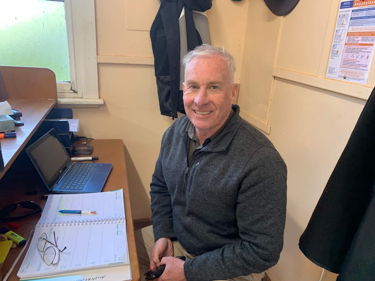 A middle aged man sits at a desk with papers and a laptop before him