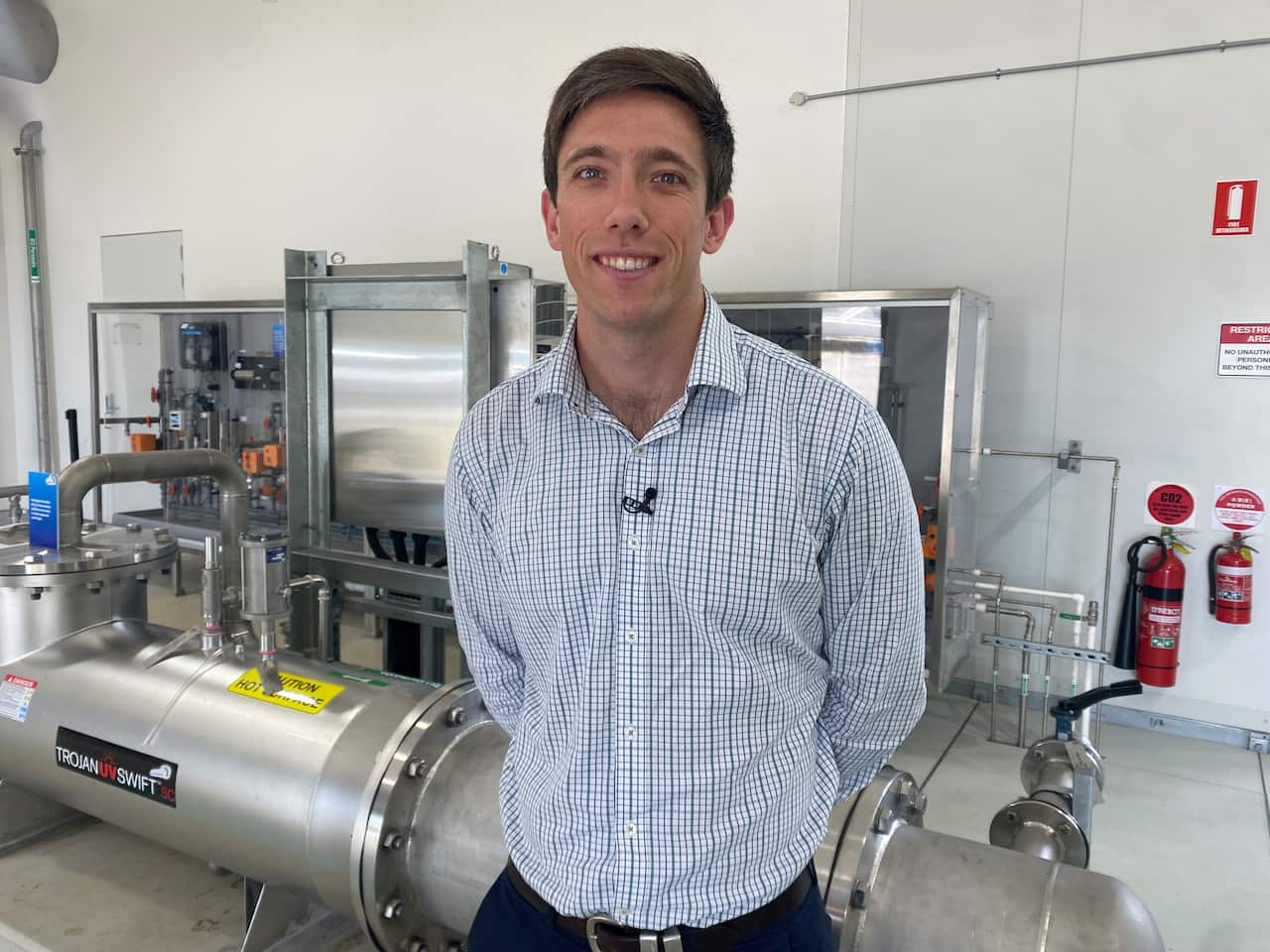 A man in his late 30s, wearing a chequered blue and white shirt and standing in front of large metal pipes.