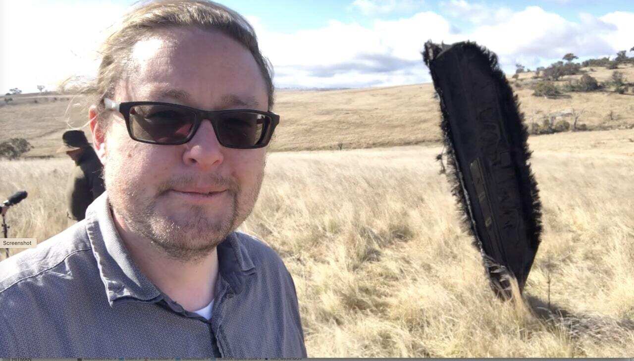 A man wearing glasses standing in a field in front of a large metal item sticking out of the ground.