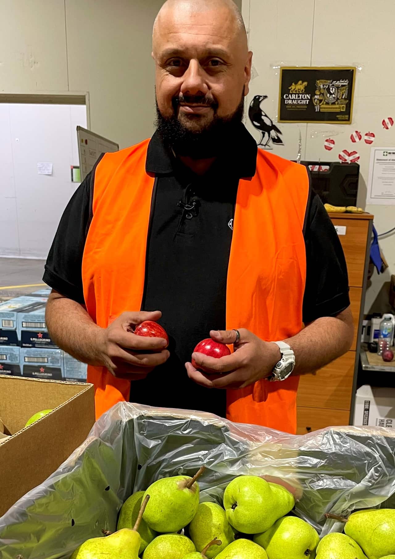 A man in a hi viz vest holding apples and standing in front of  box of pears.