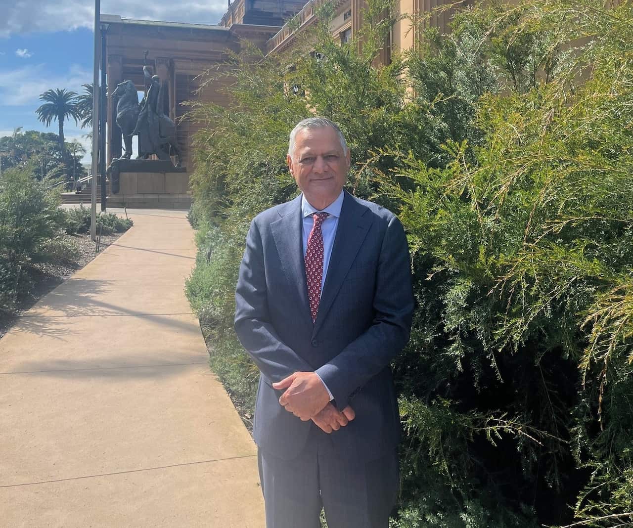 A man in a suit is smiling as he poses next to a bush outside.