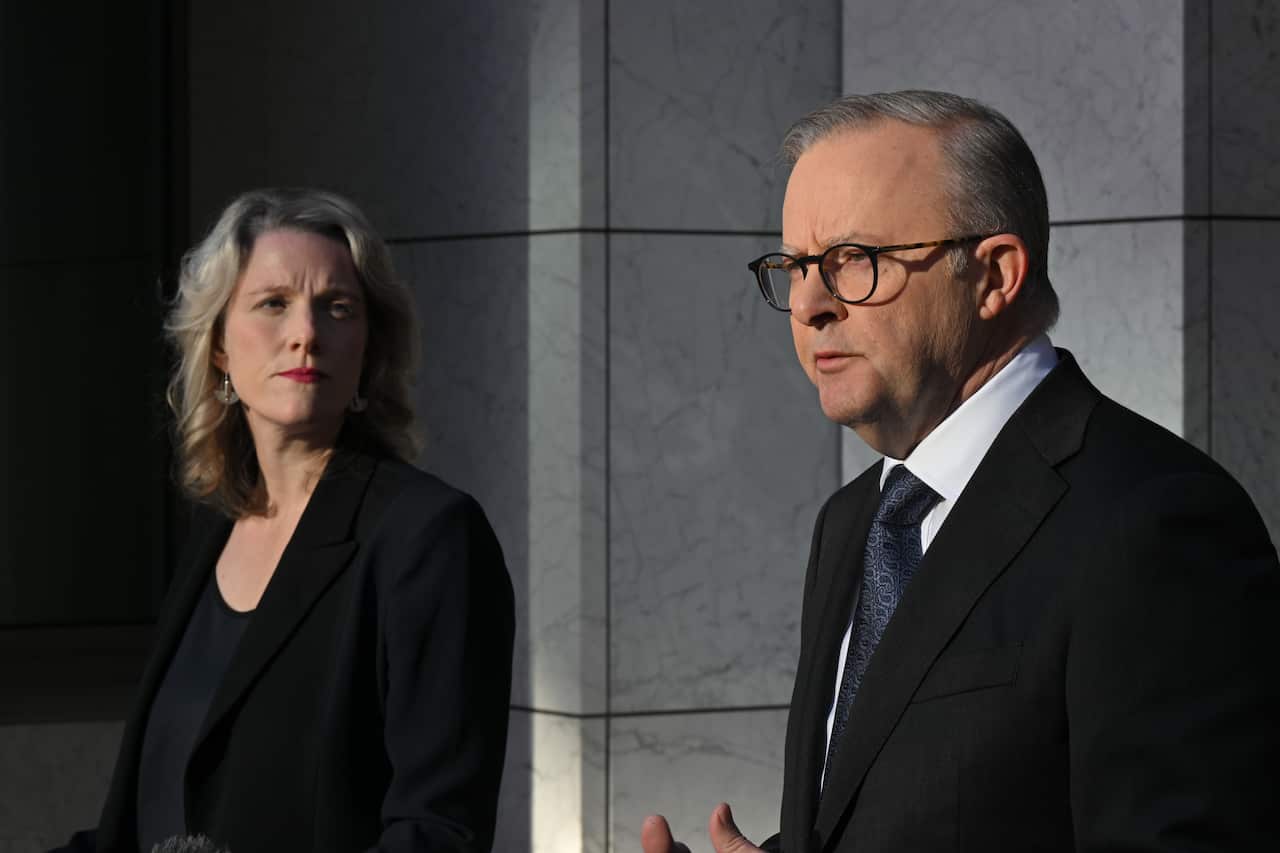 A man wearing a suit, tie and glasses speaks while a woman wearing a dark jacket and top