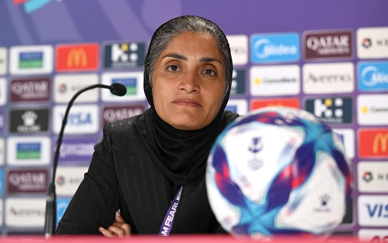A woman in a black head scarf with grey hair looks directly at the lens with a football in front of her