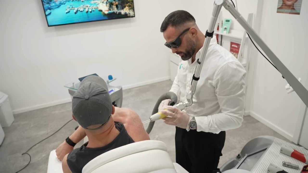 A man in a white shirt and protective glasses points a laser at a client wearing a black singlet sitting on a chair inside a clinic.
