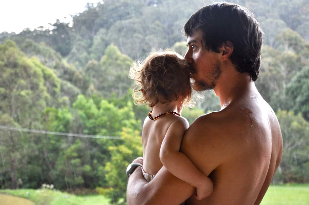 A bare-chested father and child, him holding and kissing her head as she looks out at the forest.