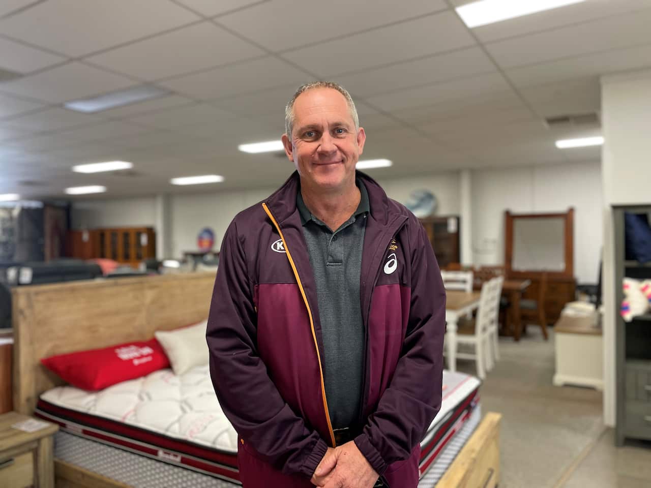 Brett stands and smiles in front of a bed and other furniture on display in his store.