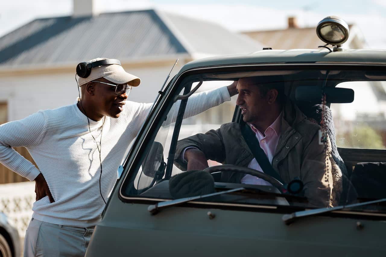 A young man with headphones perched on his cap leans on the side of a vehicle. Another man sits inside the vehicle. 