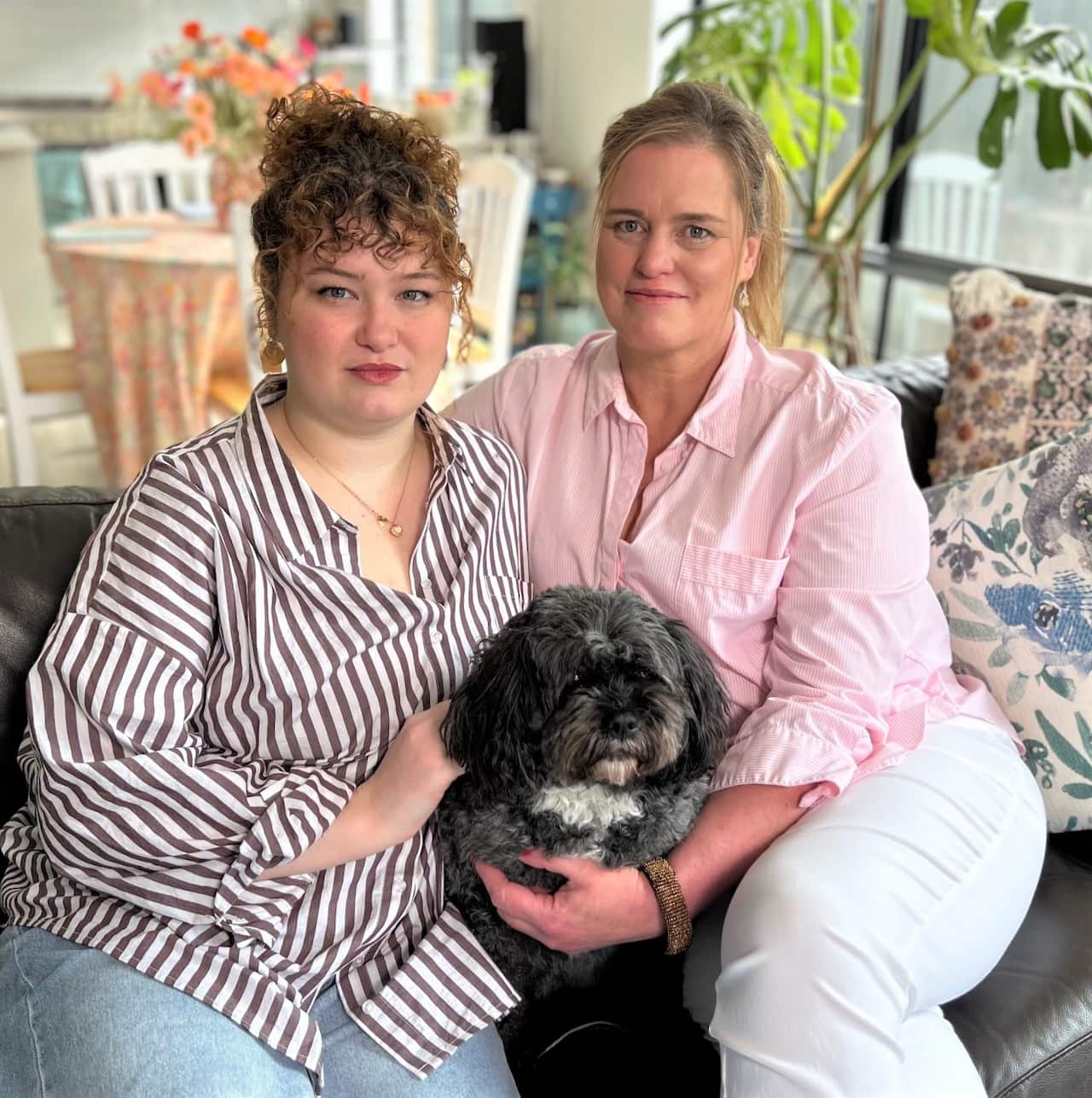 A woman in a striped shirt sits on a sofa with an older woman in a pink shirt holding a black dog.