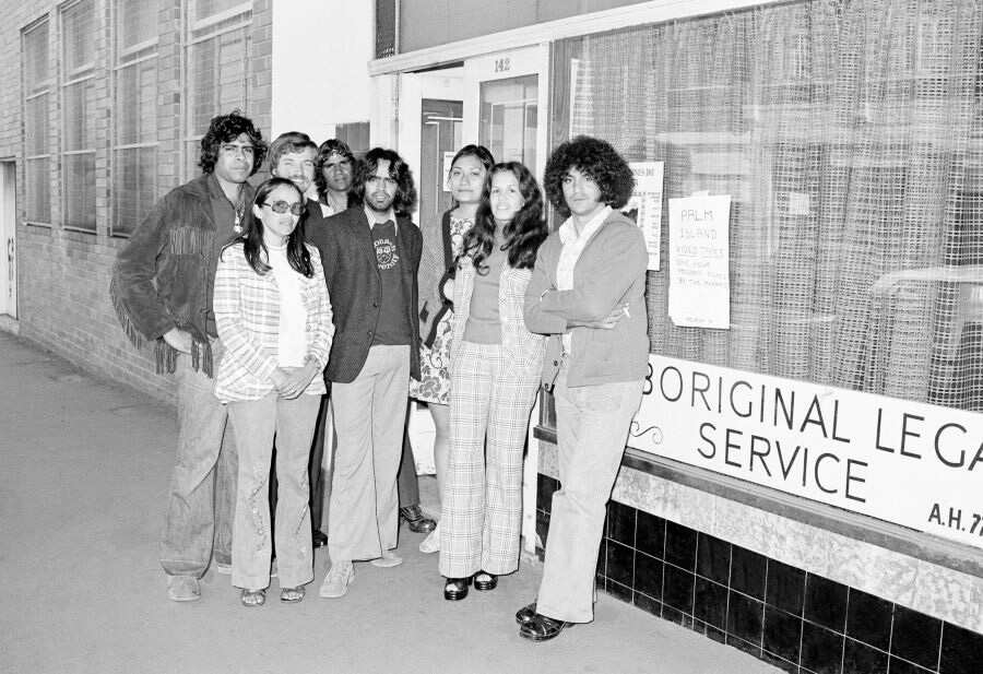 black and white photo of nine people, a mix of Blak and white, standing in front of a shop bearing the sign "Aboriginal legal service"